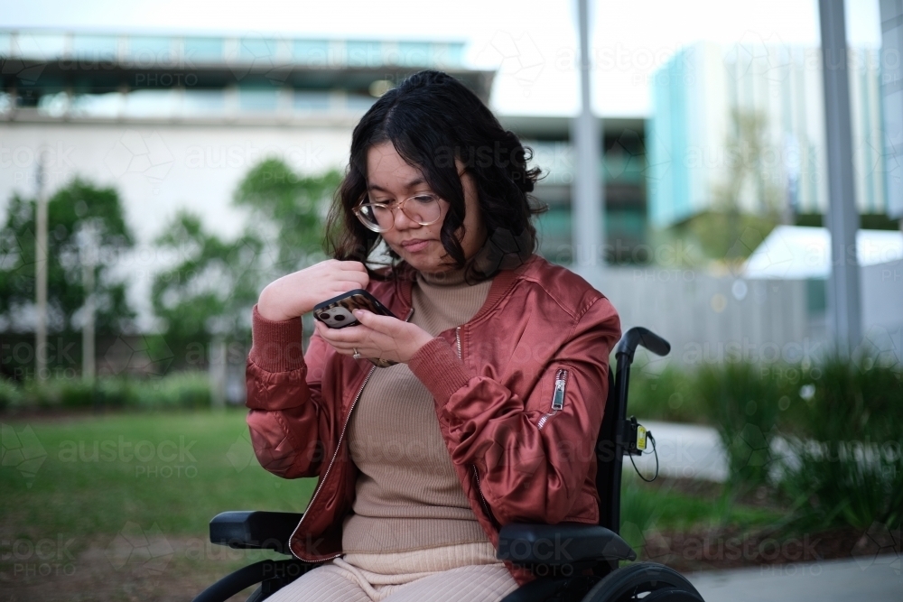 Woman with a disability sitting in a wheelchair looking down at her mobile phone - Australian Stock Image