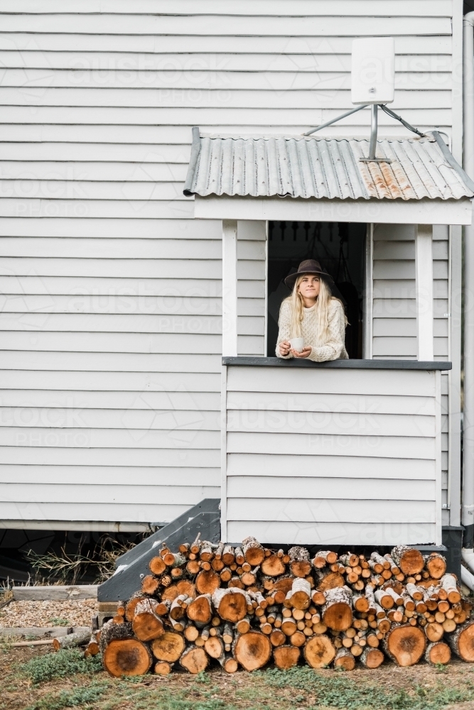 Woman with coffee standing at entrance to weatherboard home behind pile of firewood - Australian Stock Image
