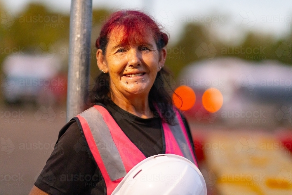 woman with coloured hair and face piercings in hi-vis vest holding a hard hat with blurry background - Australian Stock Image