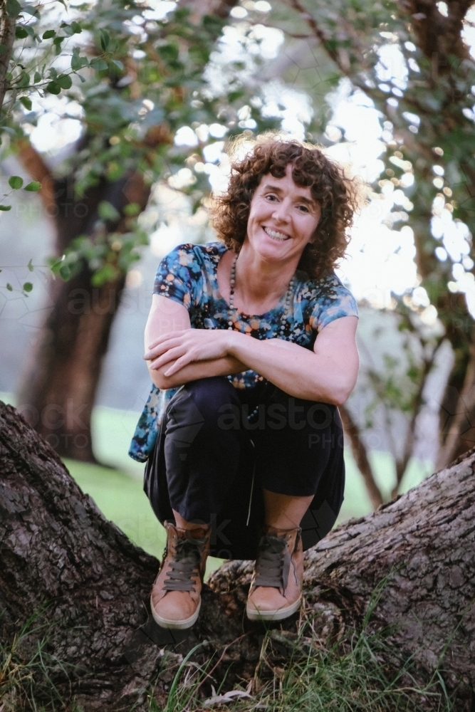 woman with curly hair and casually dressed sitting on tree trunk - Australian Stock Image