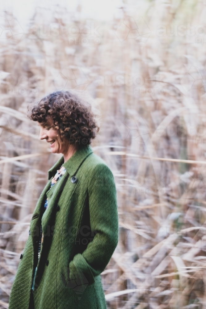 Woman with curly hair posing in front of bushland and reeds grass wearing green trench coat - Australian Stock Image