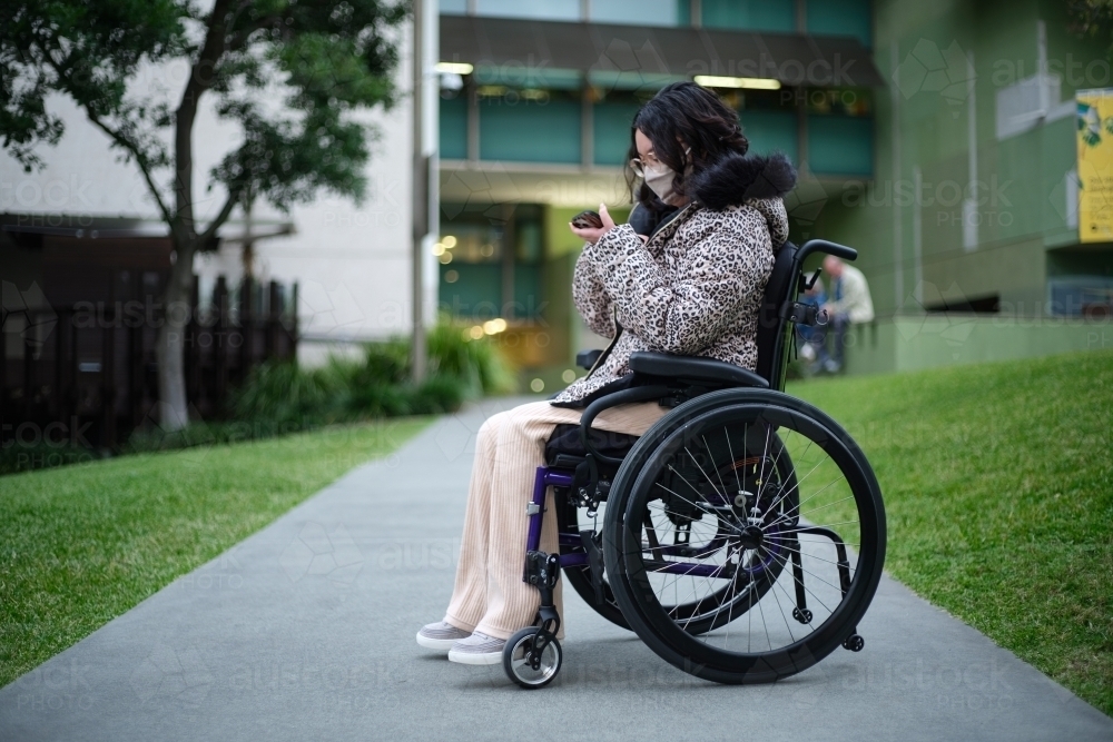 Woman with disability in a wheelchair outside looking at her mobile phone with face mask - Australian Stock Image
