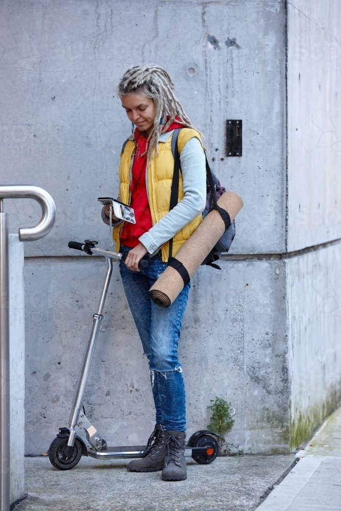 Woman with dreadlocks on electric scooter going to yoga class with phone - Australian Stock Image