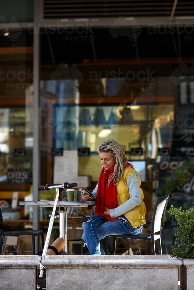 Woman with dreadlocks texting at cafe with electric scooter - Australian Stock Image
