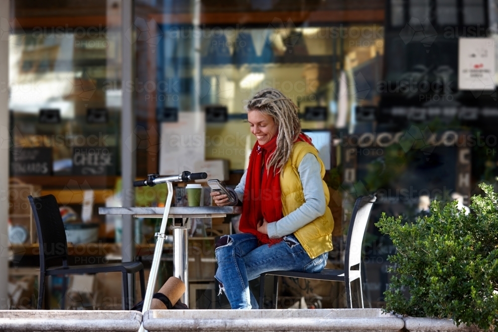 Woman with dreadlocks using her mobile phone at cafe with electric scooter - Australian Stock Image