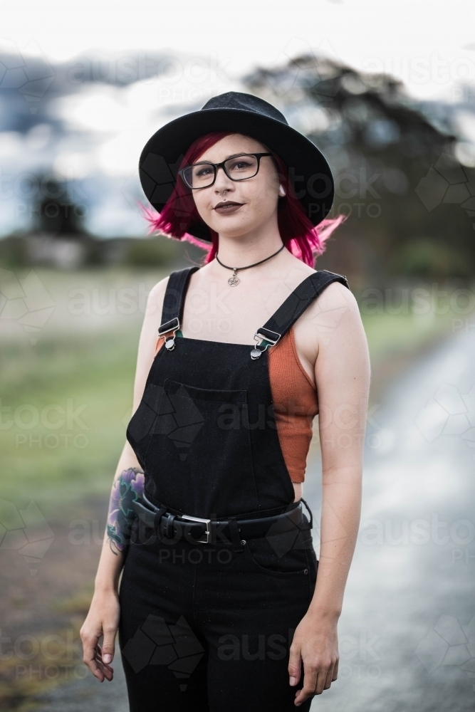 Woman with glasses and hat standing while wind blows pink hair - Australian Stock Image