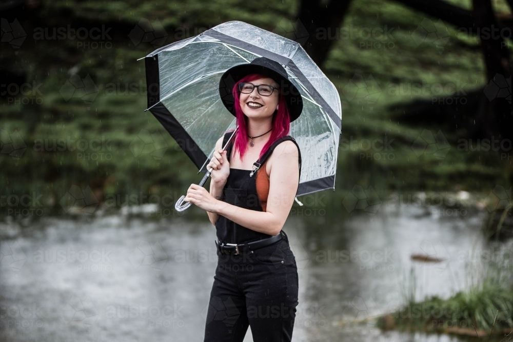Woman with pink hair standing in rain holding umbrella near river - Australian Stock Image