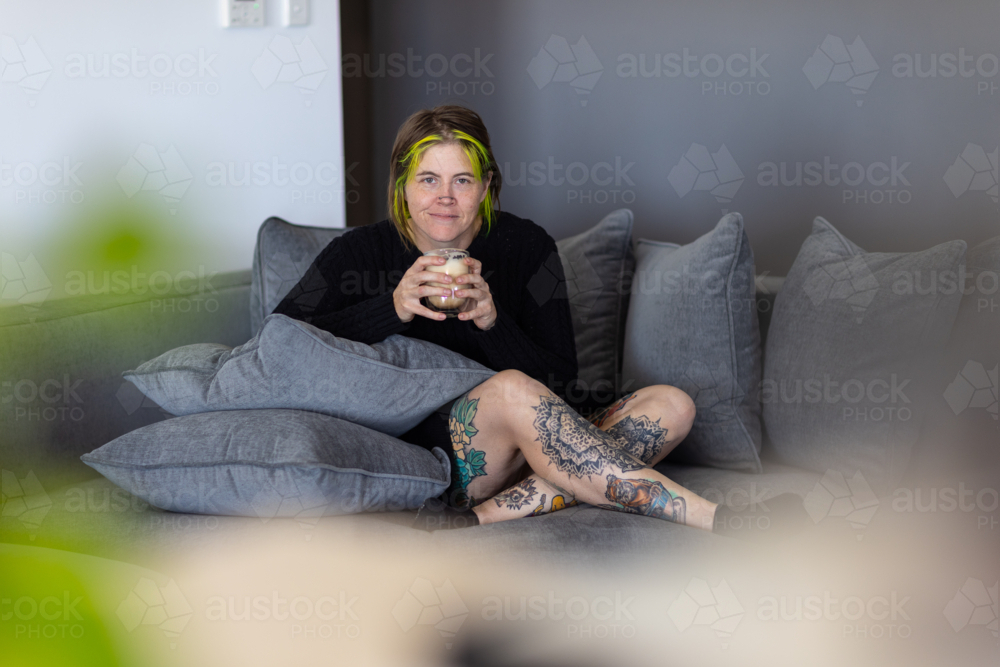 woman with tattoos, dyed hair and nose chain sitting cross-legged on sofa with cup of coffee - Australian Stock Image