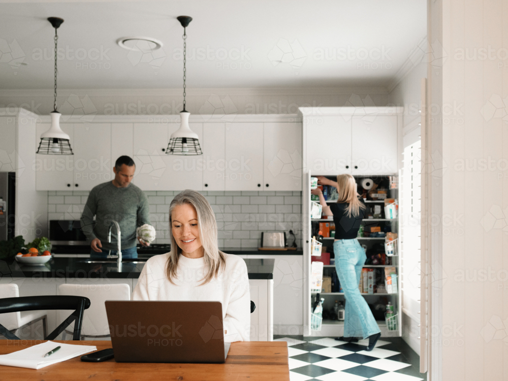 Woman working from home on her laptop at wooden table while husband works in the kitchen - Australian Stock Image