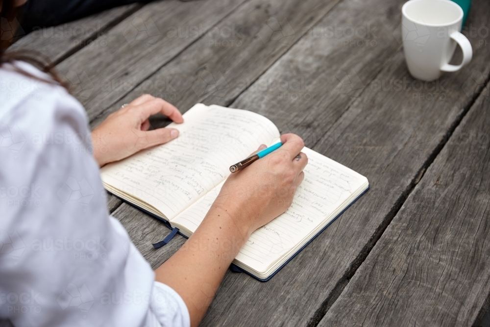 Woman writing on notepad on wooden table outdoors - Australian Stock Image