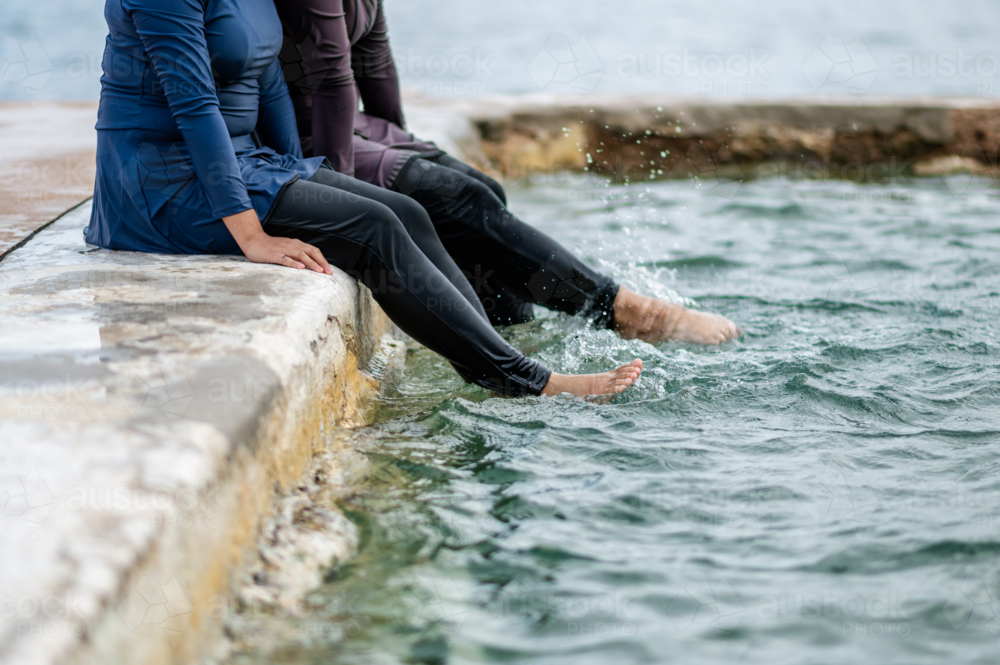 Women in modest swimwear sitting on stone platform kicking their legs in the tidal pool - Australian Stock Image