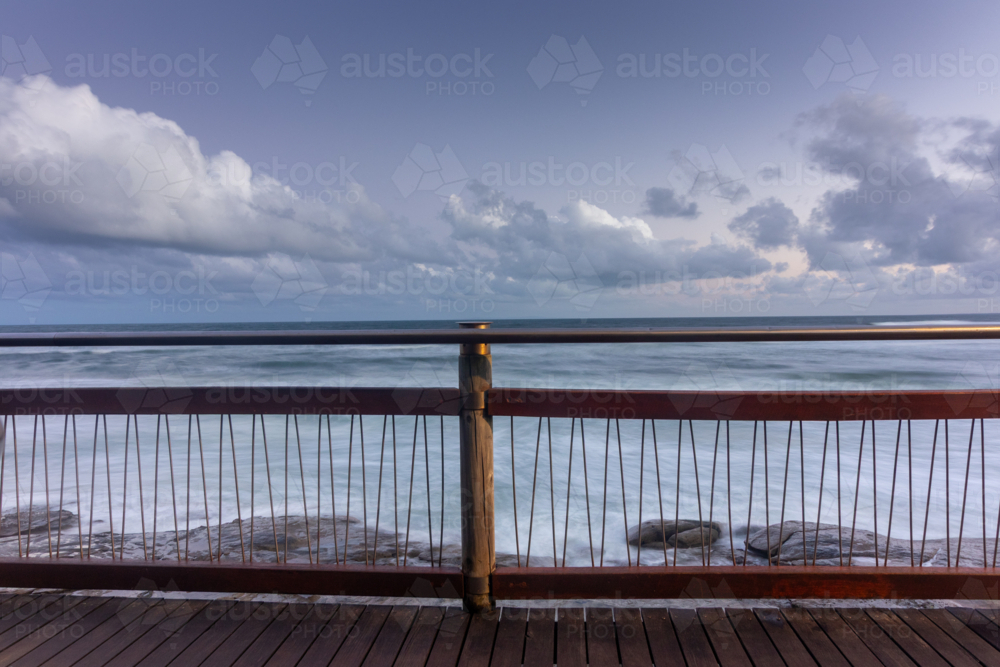 Wooden boardwalk and metal railing overlooking rocks and ocean with cloudy sky - Australian Stock Image