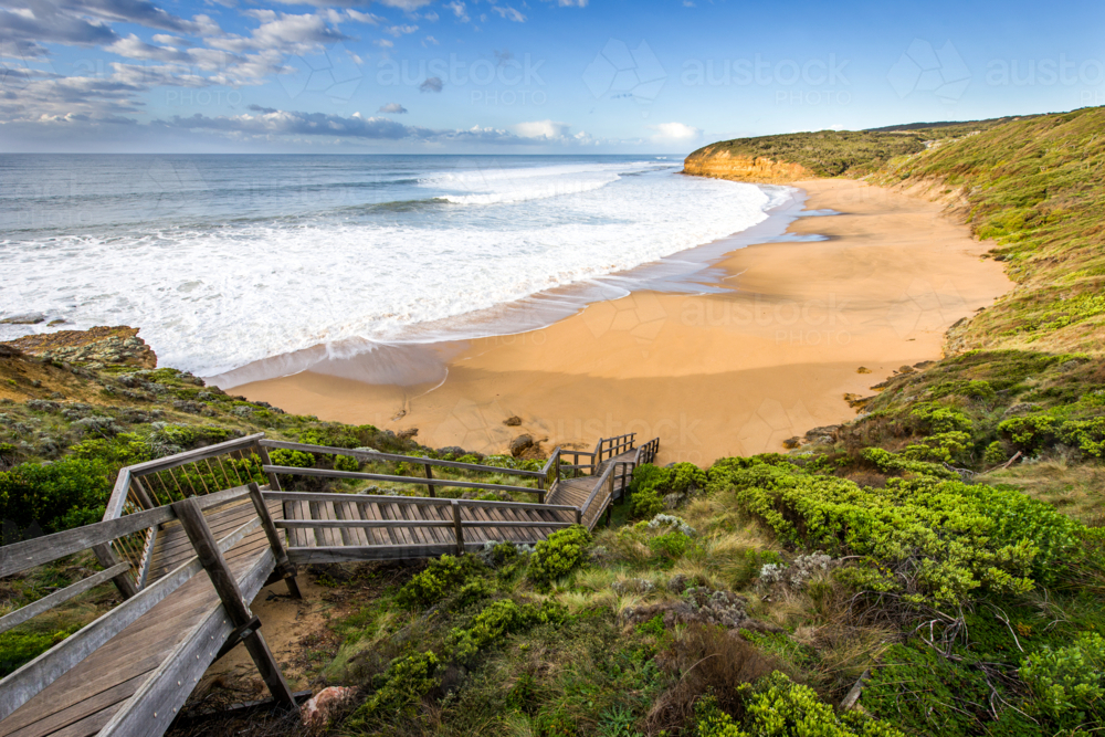 Wooden staircase leading down to a sandy beach with rolling waves and rugged cliffs - Australian Stock Image