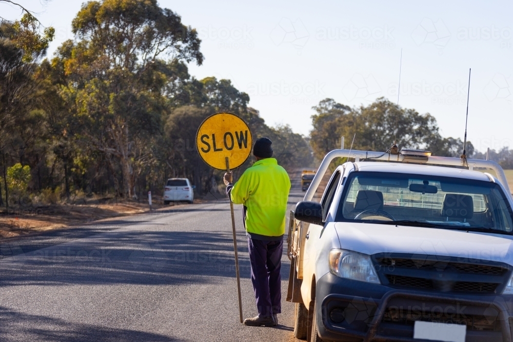 worker holding slow lollipop sign on edge of road near vehicle - Australian Stock Image