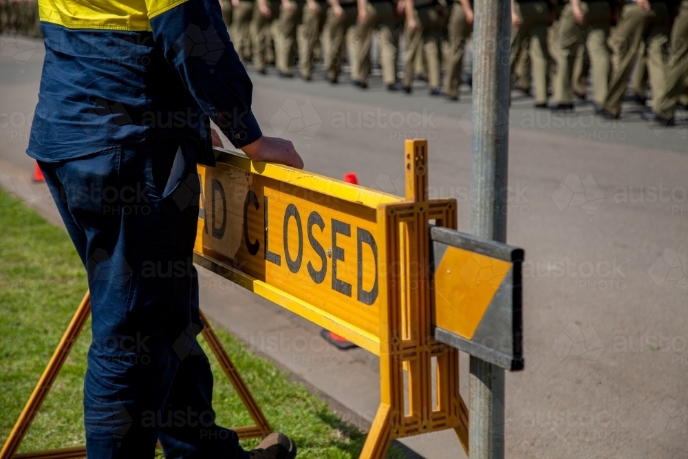 Worker in yellow clothing leaning on a road closed sign while soldiers march past - Australian Stock Image