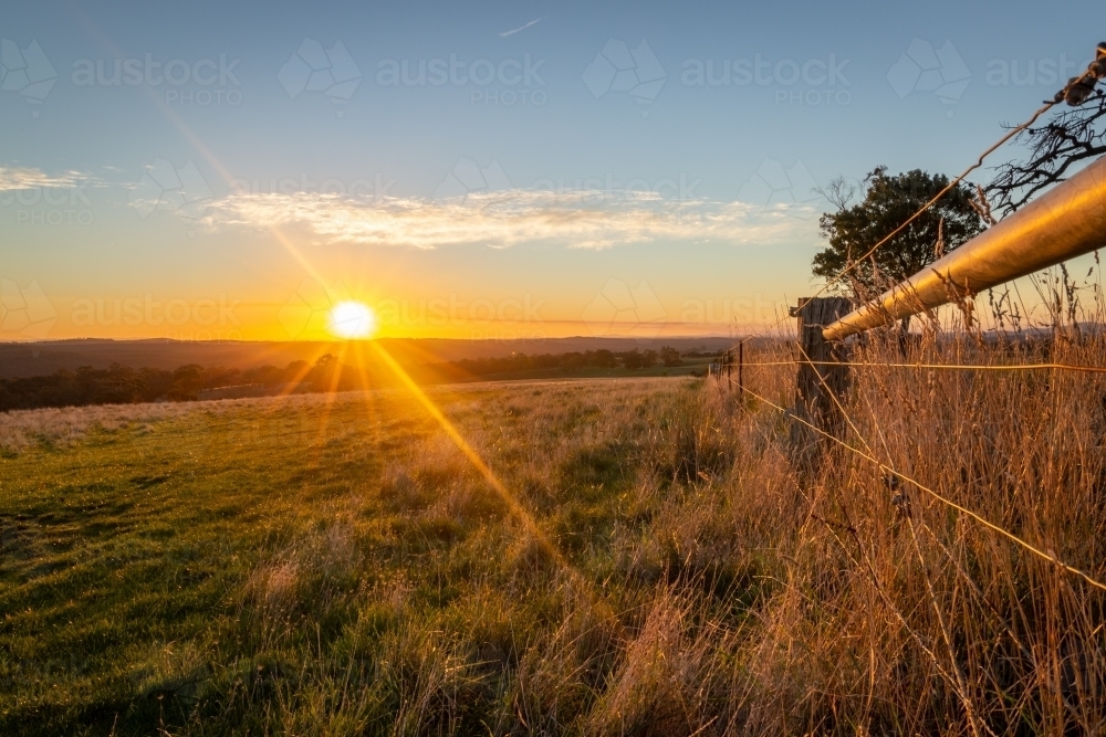 Yellow Sun setting in the distance over hills and paddocks with a farm fence framing the image - Australian Stock Image