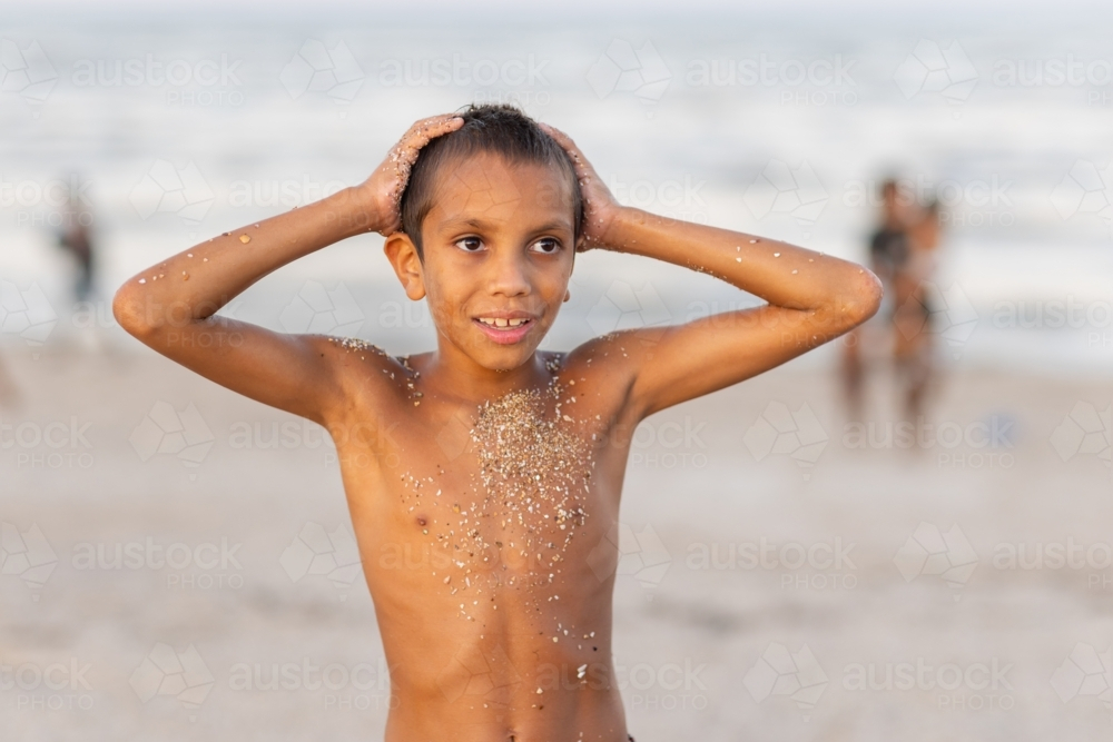 young aboriginal boy shirtless with hands on head on beach with coarse sand sticking to his skin - Australian Stock Image