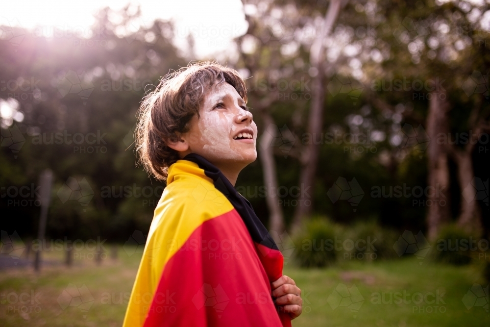 Young Aboriginal boy wearing Aboriginal flag around his shoulders and looking up - Australian Stock Image