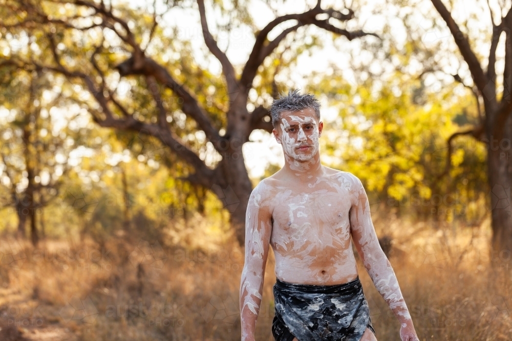 Young aboriginal man wearing ochre body paint and a lap lap walking through australian bushland - Australian Stock Image