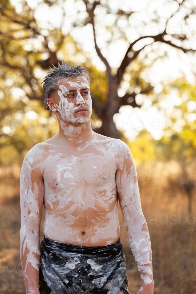 Young aboriginal man with neutral expression standing on country with ochre body paint - Australian Stock Image