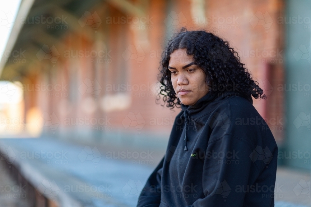 Young aboriginal woman wearing black hoody against blurred background of old station platform - Australian Stock Image
