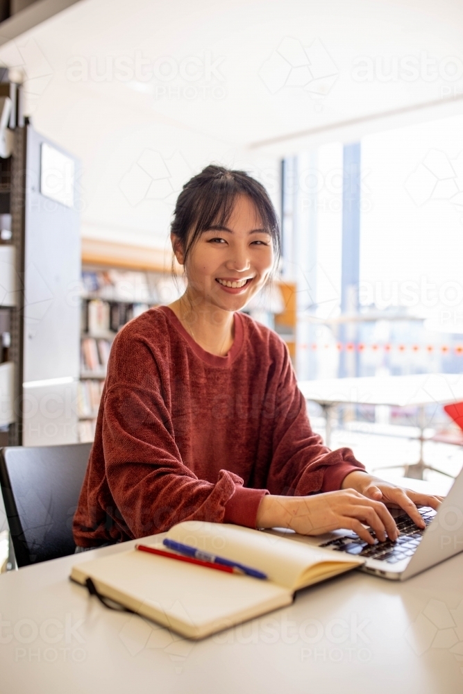 Young adult asian woman at the library studying and using her laptop - Australian Stock Image