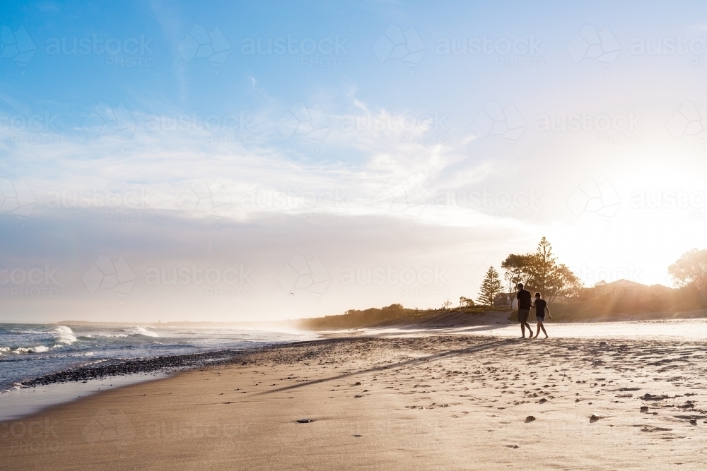 Young adult couple take a romantic walk along a pebbly beach at sunset. - Australian Stock Image