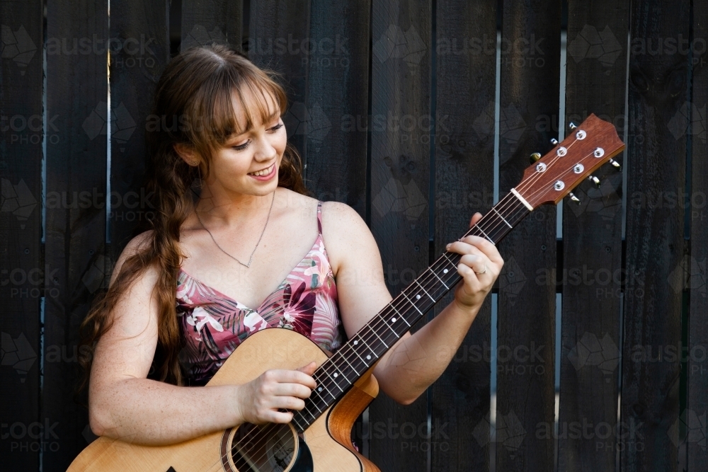 Young adult guitar player playing instrument outside leaning on black wooden fence - Australian Stock Image