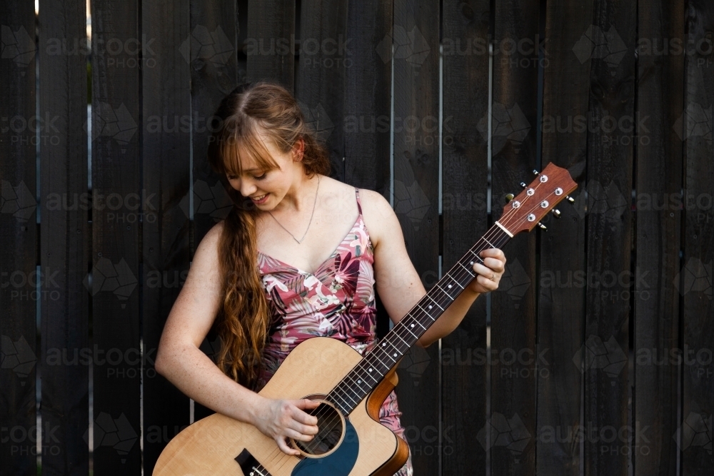 Young adult guitar player playing instrument outside leaning on black wooden fence - Australian Stock Image
