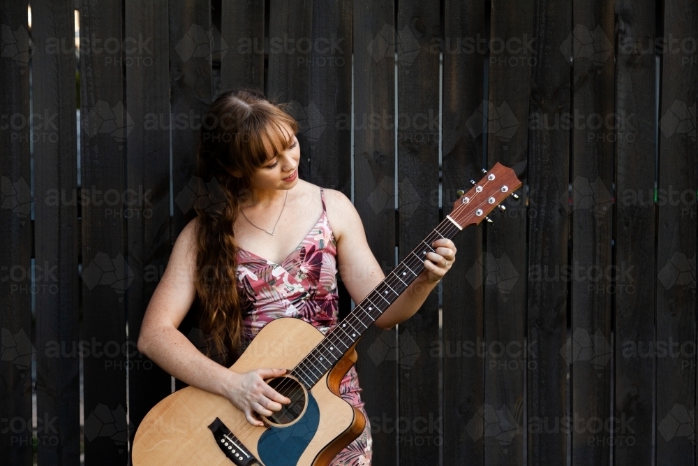 Young adult guitar player playing instrument outside leaning on black wooden fence - Australian Stock Image