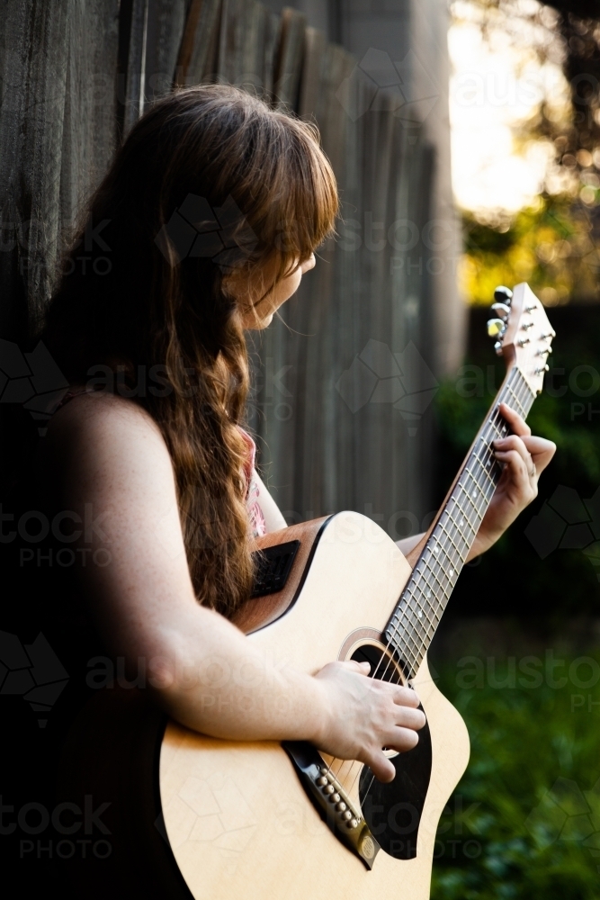 Young adult guitar player playing instrument outside leaning on black wooden fence - Australian Stock Image