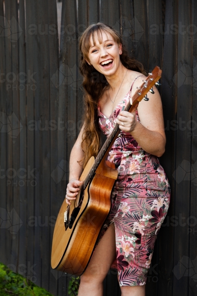 Young adult guitar player playing instrument outside leaning on black wooden fence - Australian Stock Image