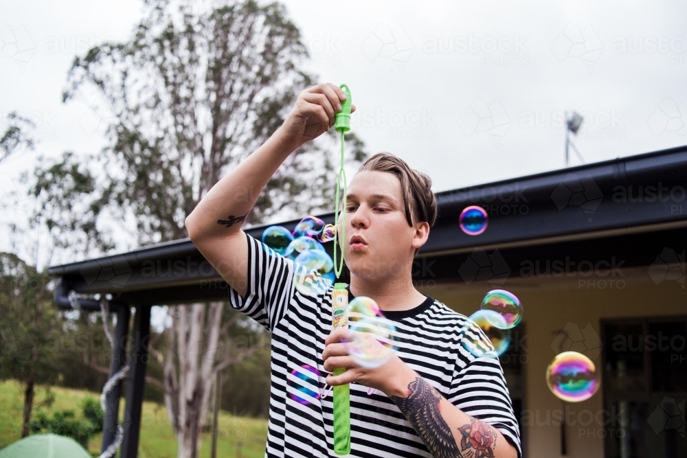 Young adult male with tattoos blowing multiple bubbles outside. - Australian Stock Image