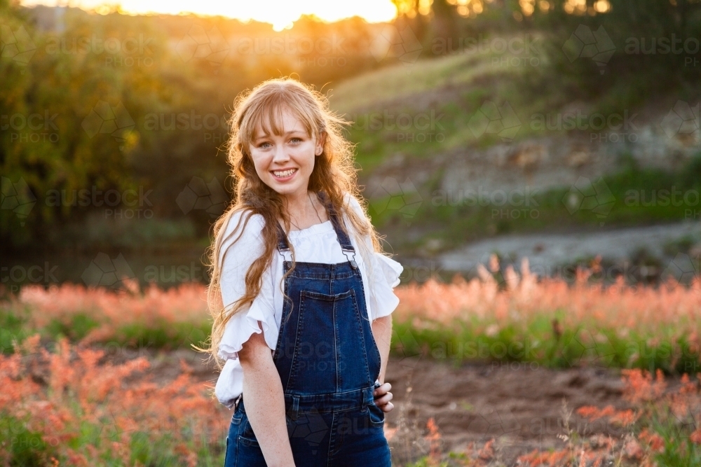 Young adult standing outside by river at sunset in overalls - Australian Stock Image