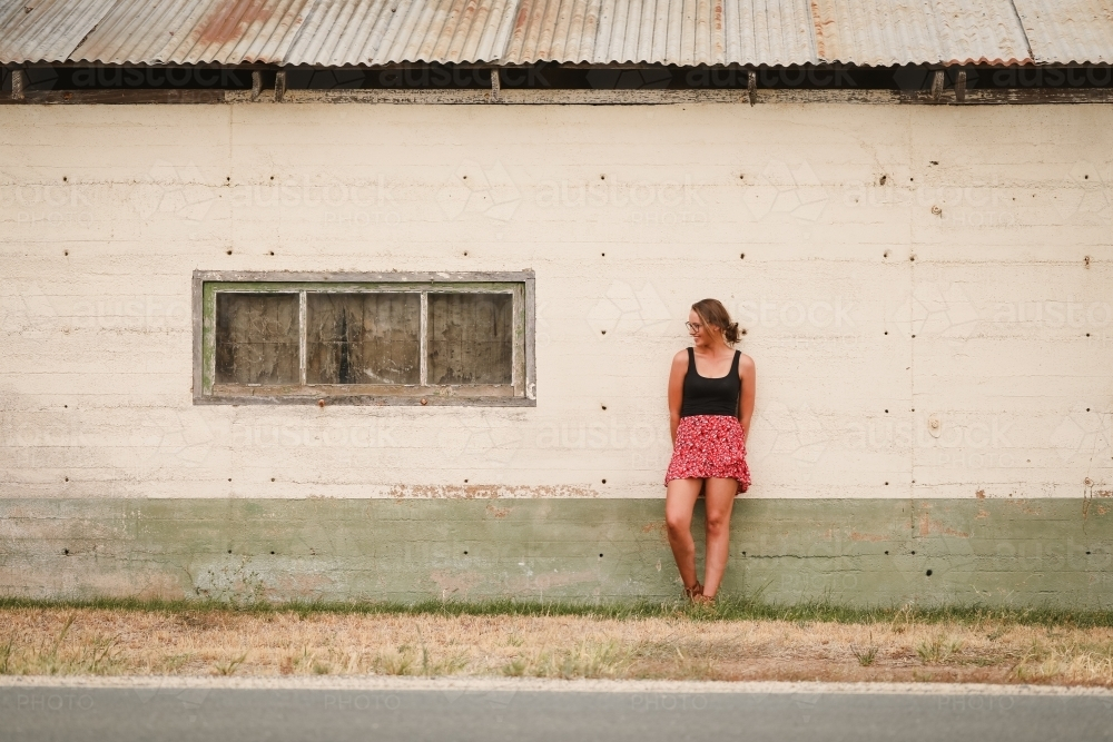 Young adult woman leaning against old building in remote farming community - Australian Stock Image