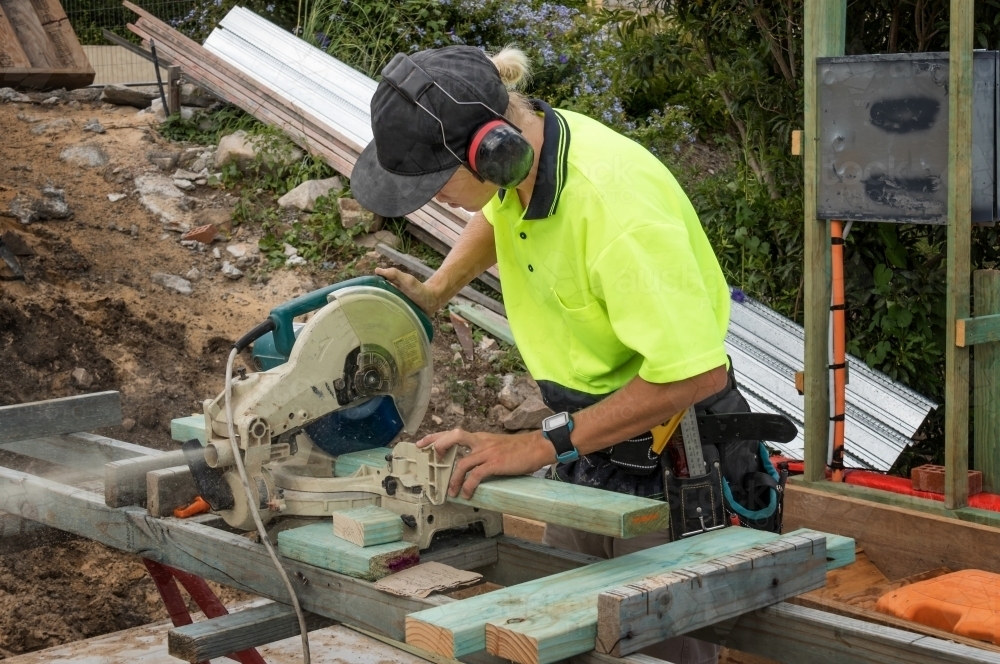 Young apprentice builder cutting length of timber with drop saw on residential construction site - Australian Stock Image