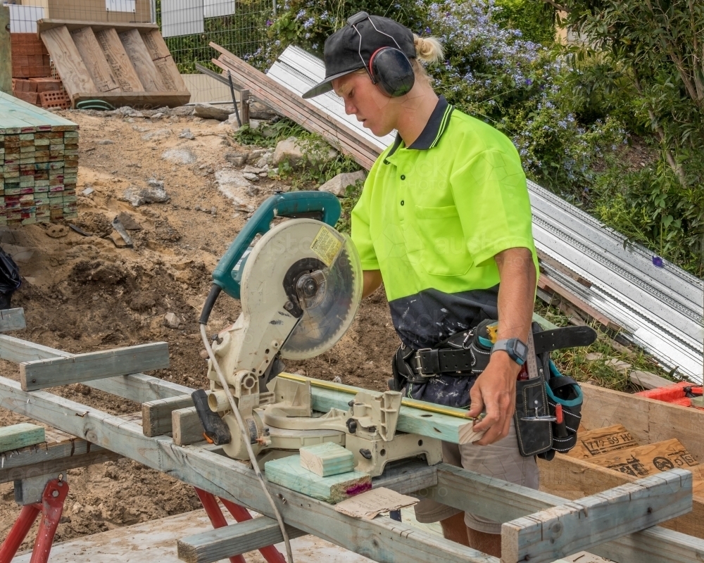 Young apprentice builder measuring and cutting timber with drop saw on residential construction site - Australian Stock Image