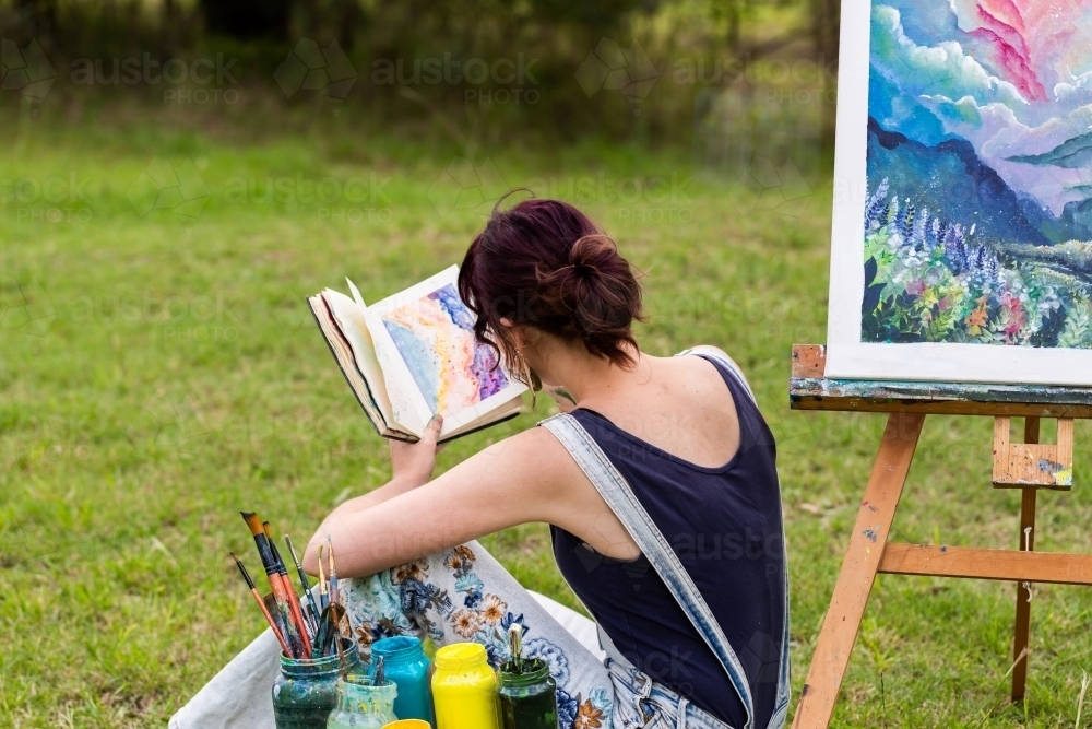 Young artist sitting beside artwork looking in notebook for inspiration - Australian Stock Image