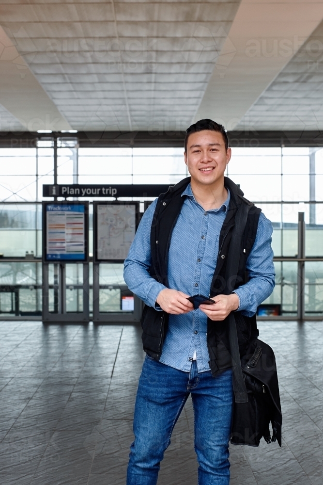 Young Asian commuter holding mobile phone waiting at train station entrance - Australian Stock Image