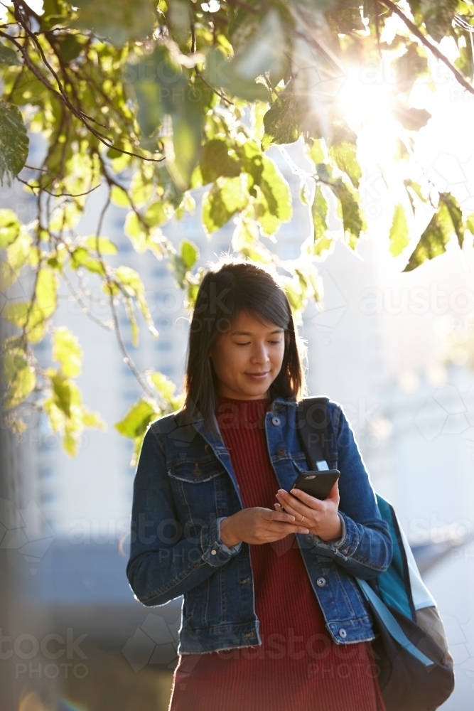 Young Asian female checking mobile phone under tree - Australian Stock Image