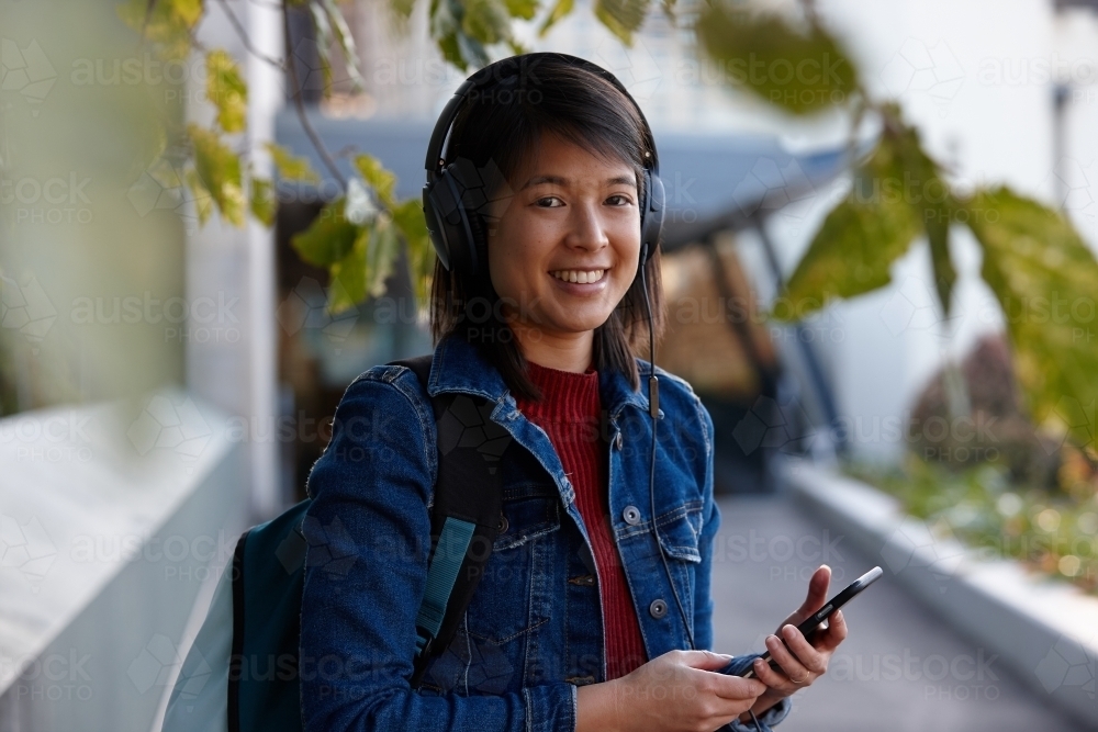 Young Asian female listening to music with headphones under tree - Australian Stock Image