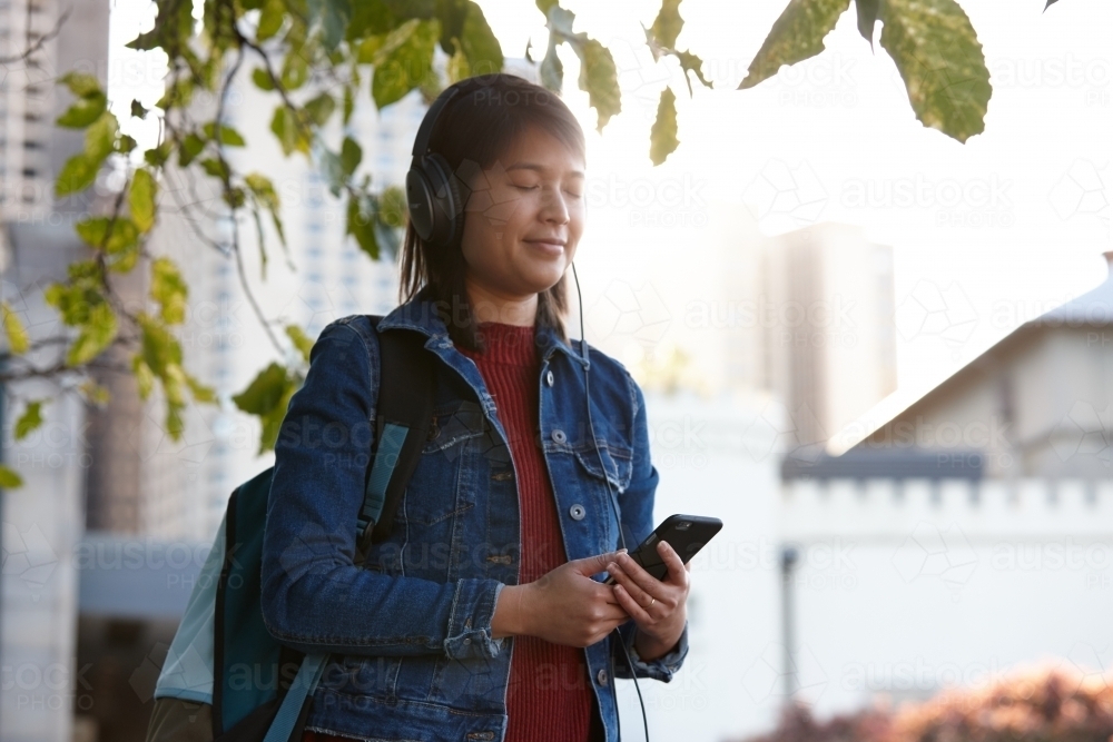 Young Asian female listening to music with headphones under tree - Australian Stock Image