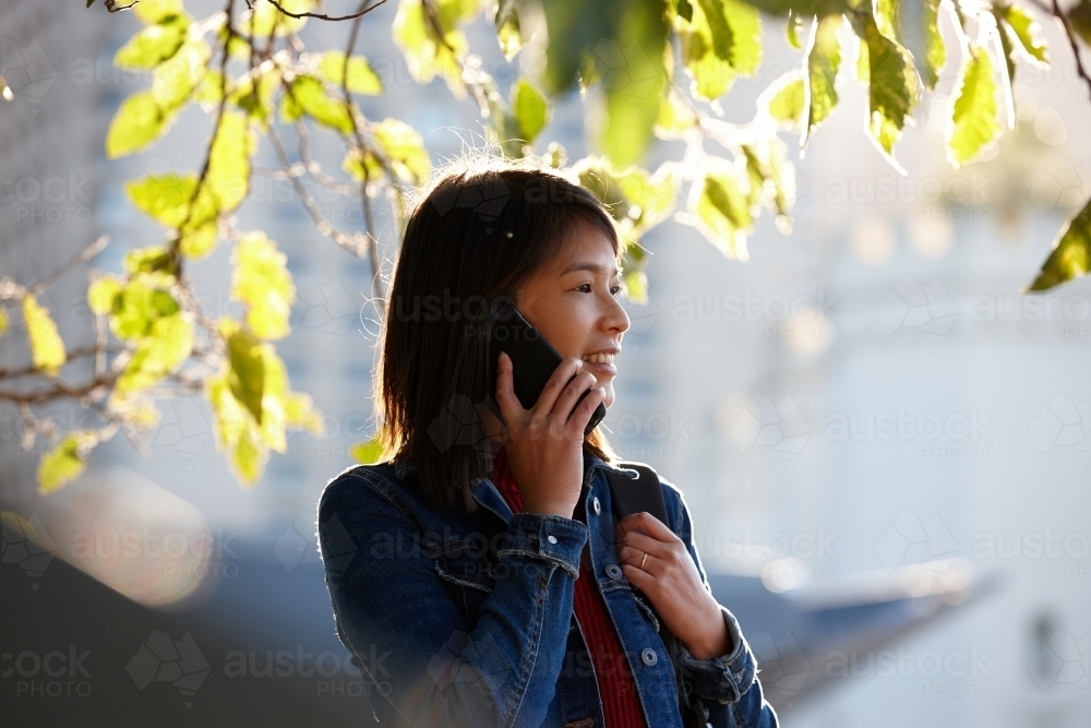 Young Asian female talking on mobile phone under tree - Australian Stock Image