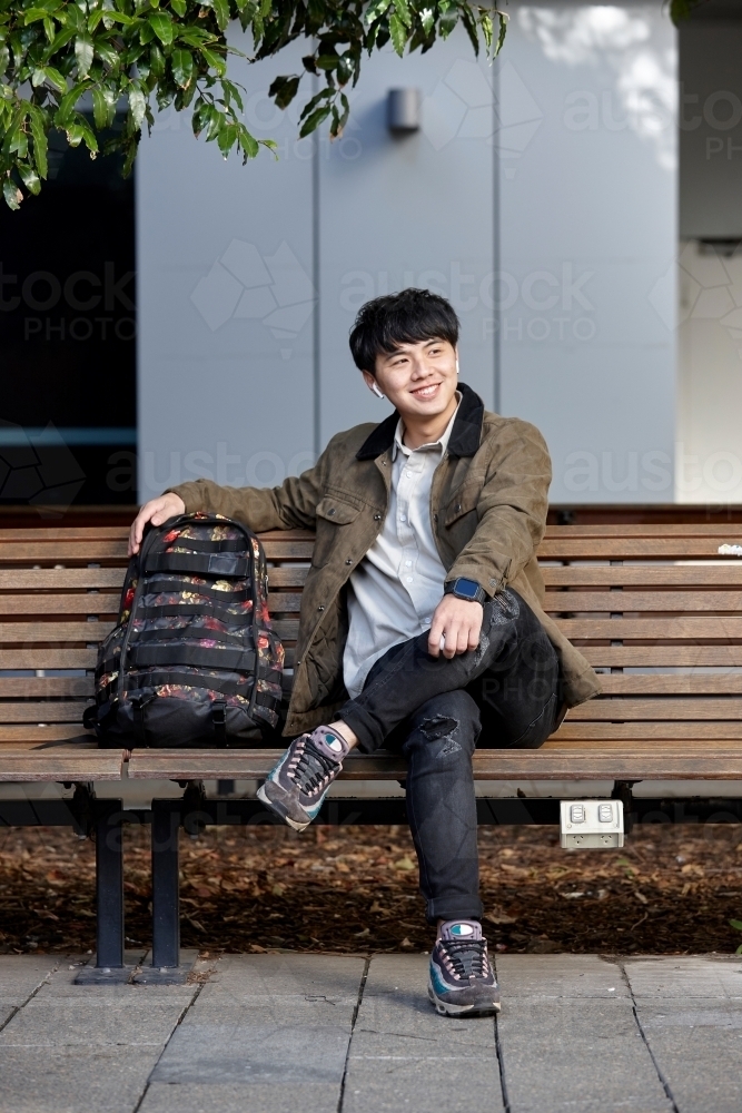 Young Asian man smiling sitting on park bench whilst listening to wireless headphones - Australian Stock Image