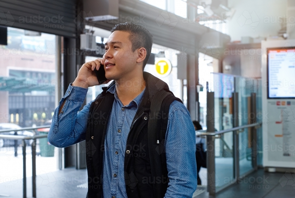 Young Asian man talking on mobile phone at train station entrance - Australian Stock Image