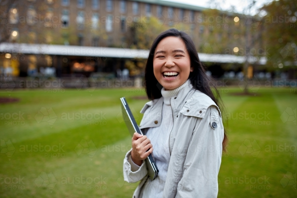 Young Asian student holding her laptop on lawn at university campus - Australian Stock Image