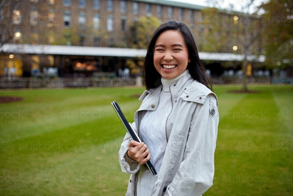 Young Asian student holding her laptop on lawn at university campus - Australian Stock Image