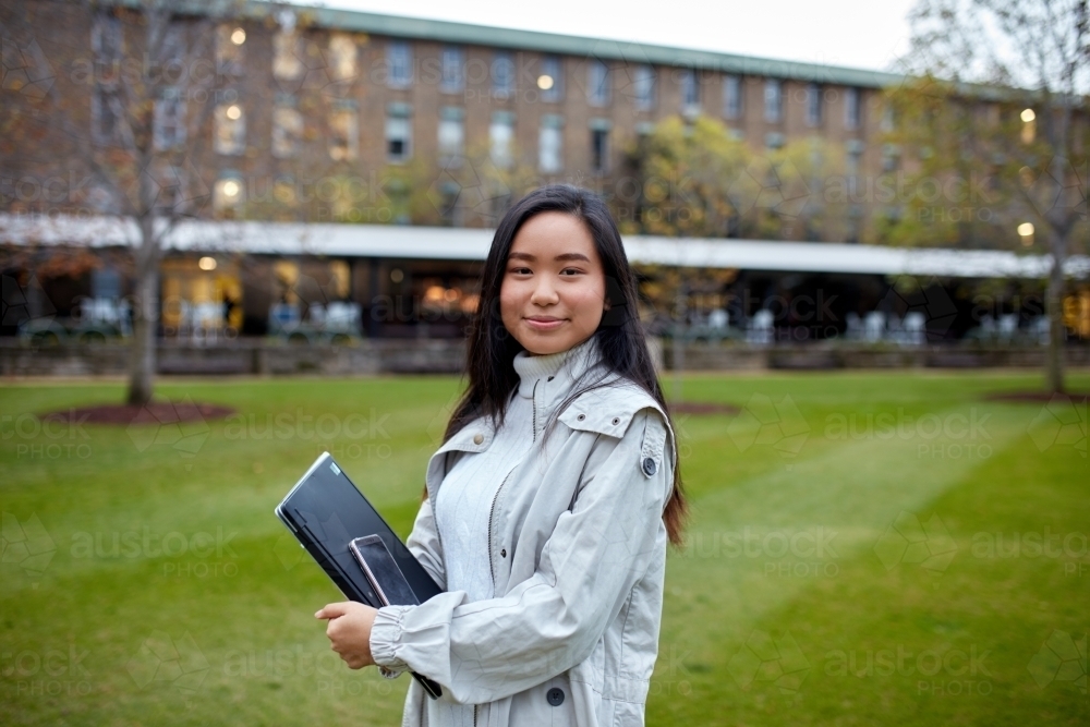 Young Asian student holding her laptop on lawn at university campus - Australian Stock Image