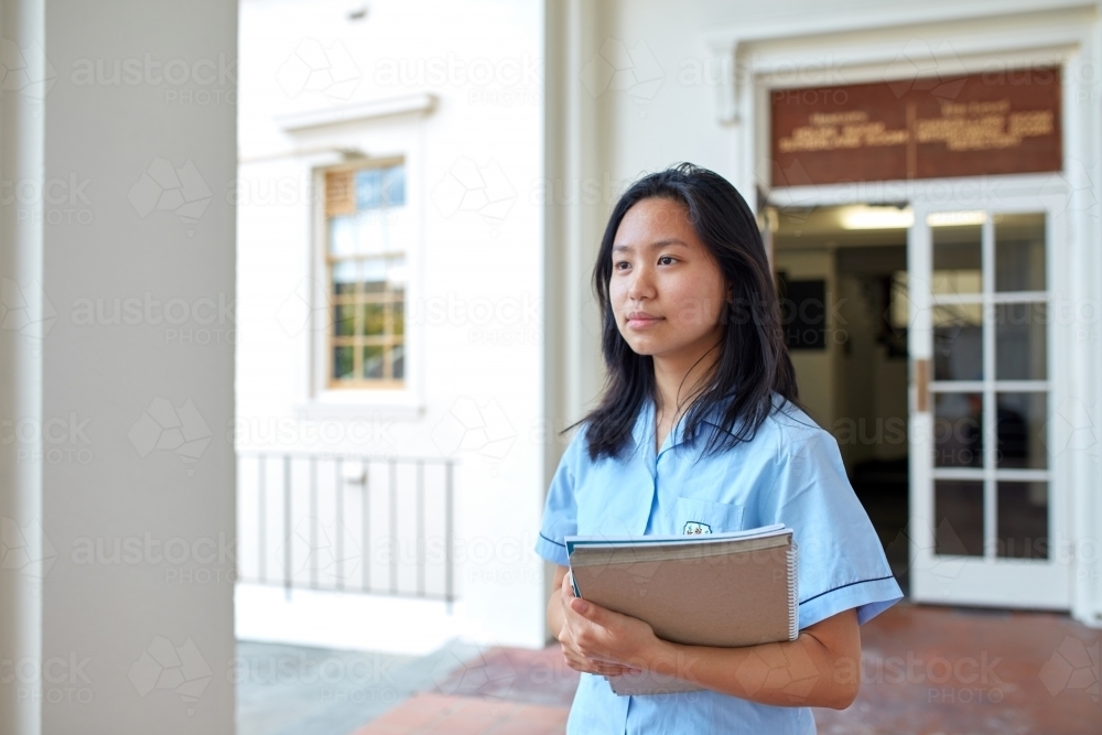 Young Asian student in front of school building holding books - Australian Stock Image