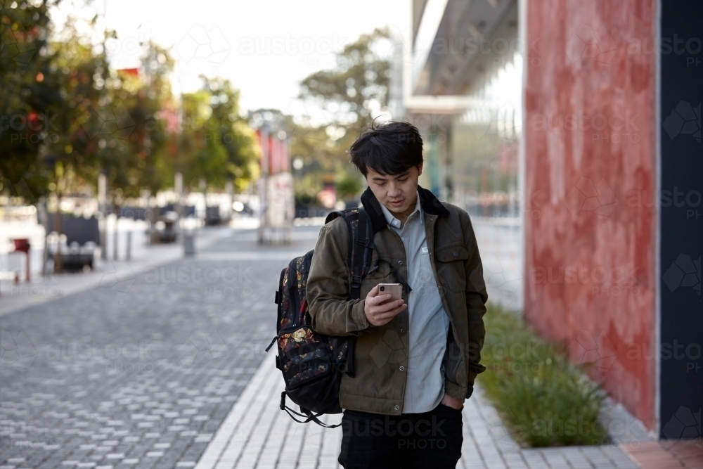 Young Asian university student walking and checking phone messages on-campus - Australian Stock Image